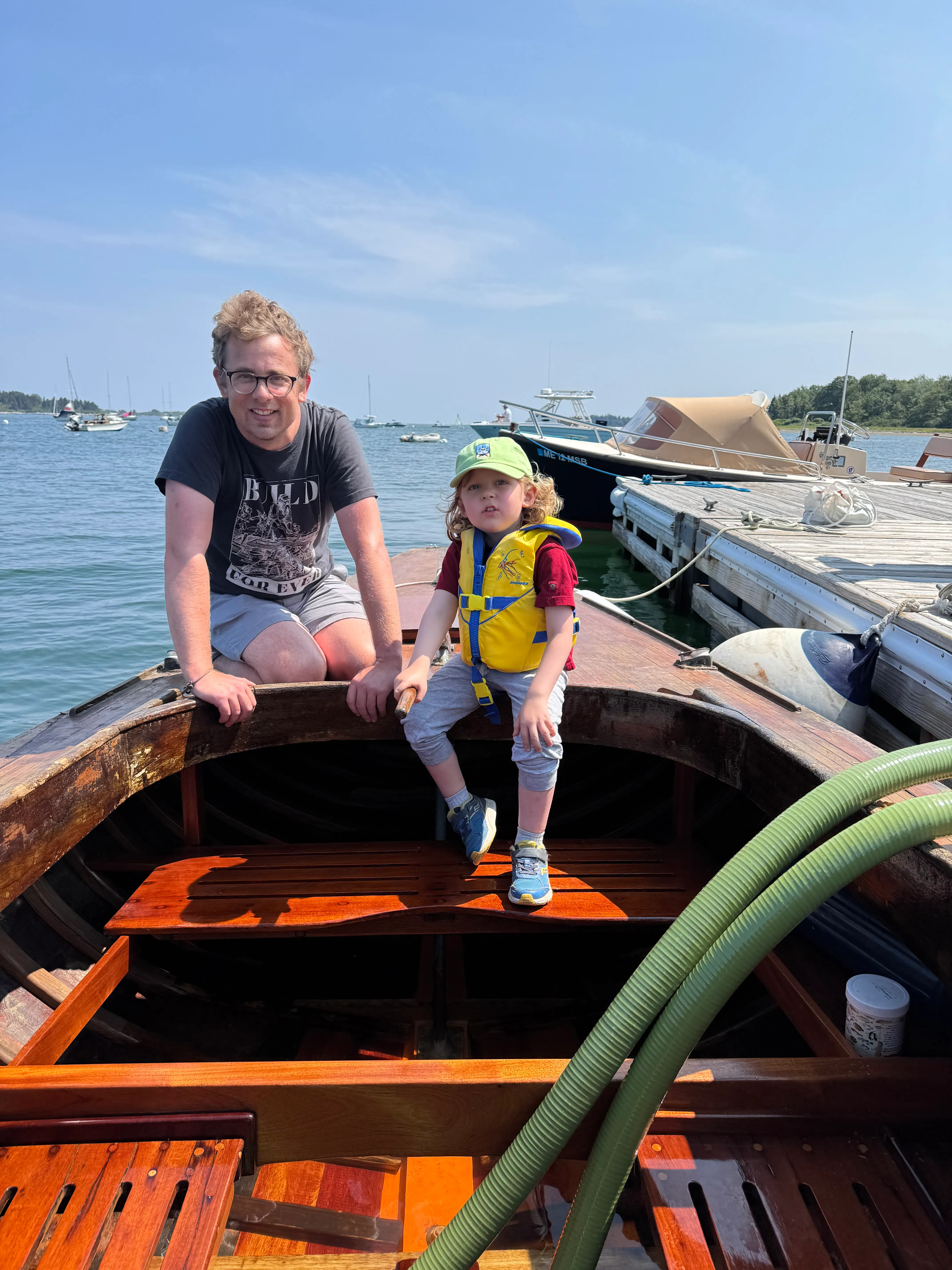 An adult and child in a yellow life jacket at the boat alongside the dock, pump hose still running