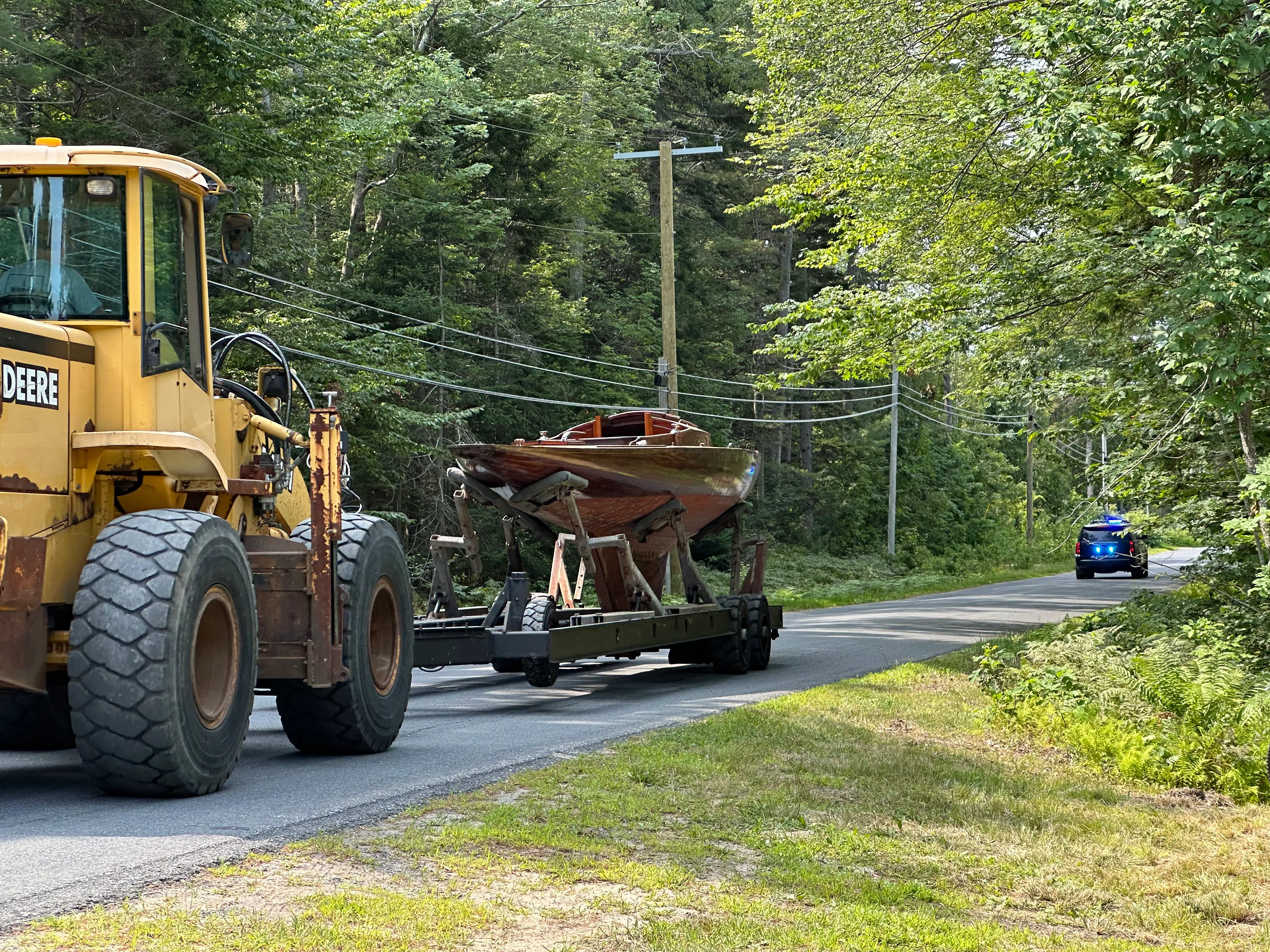 John Deere loader towing the boat down a tree-lined road with a police escort behind