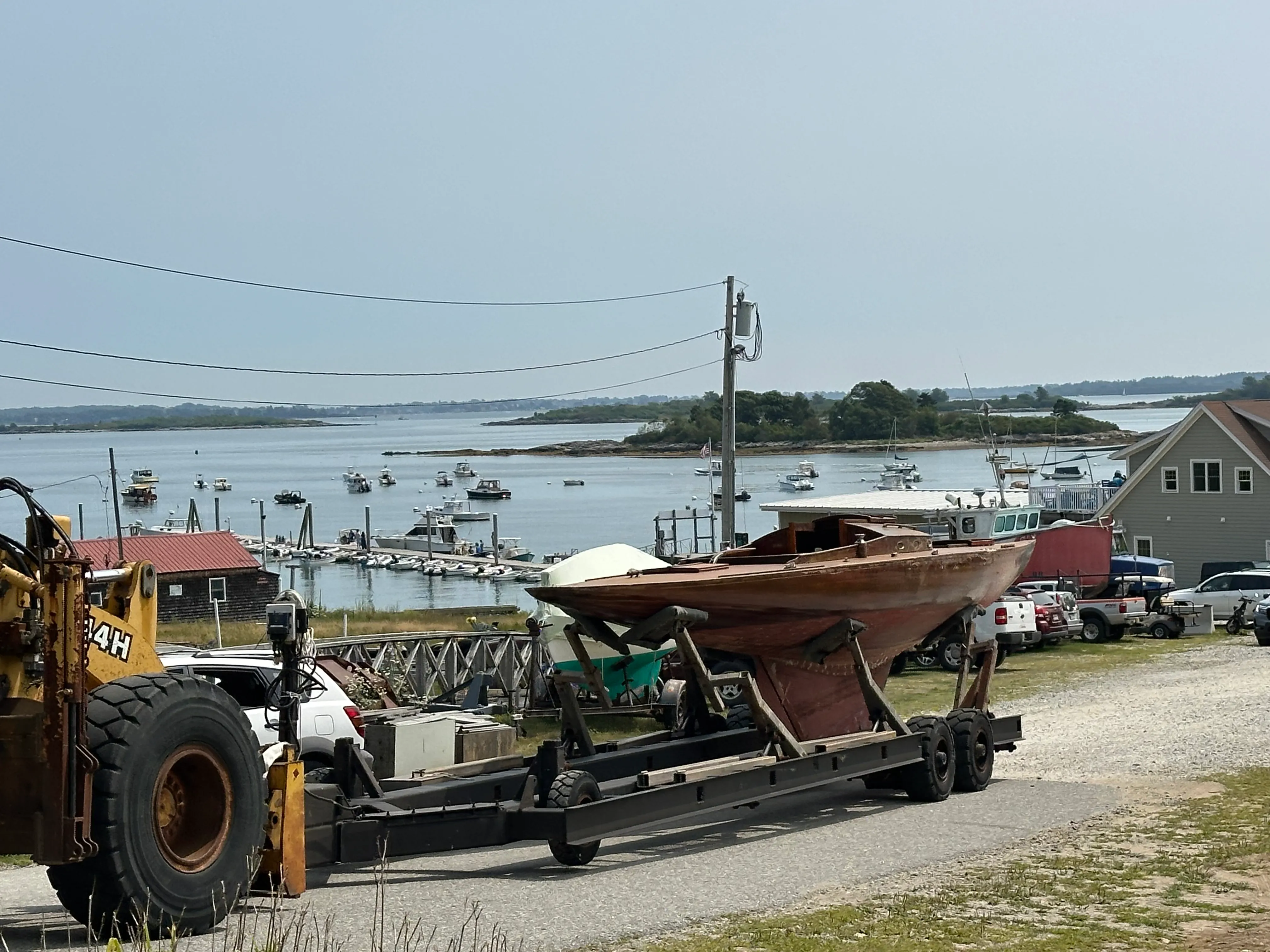 The sailboat on its trailer at the boatyard with the harbor, moored boats, and islands visible behind