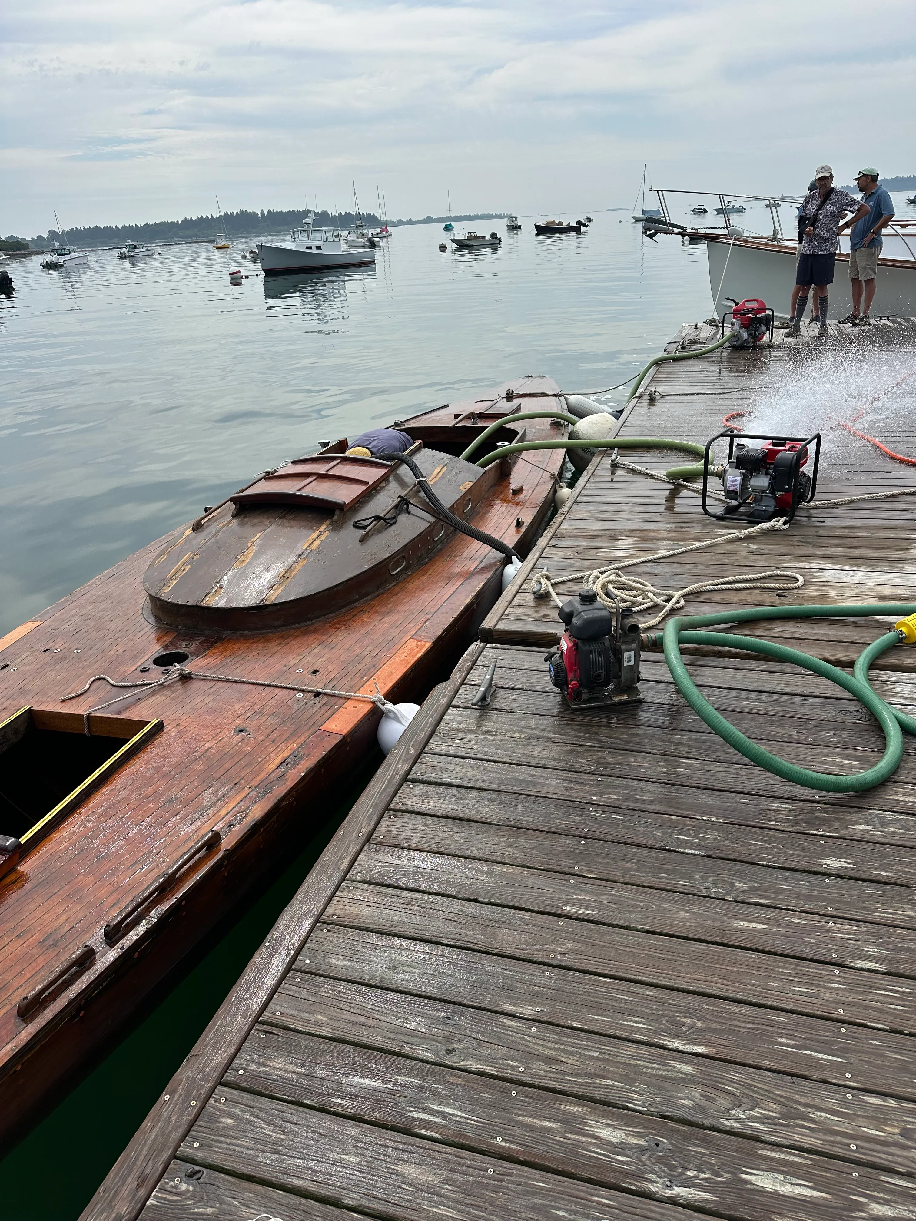 The sailboat tied alongside a dock viewed from above, with a pump and green hose on the dock