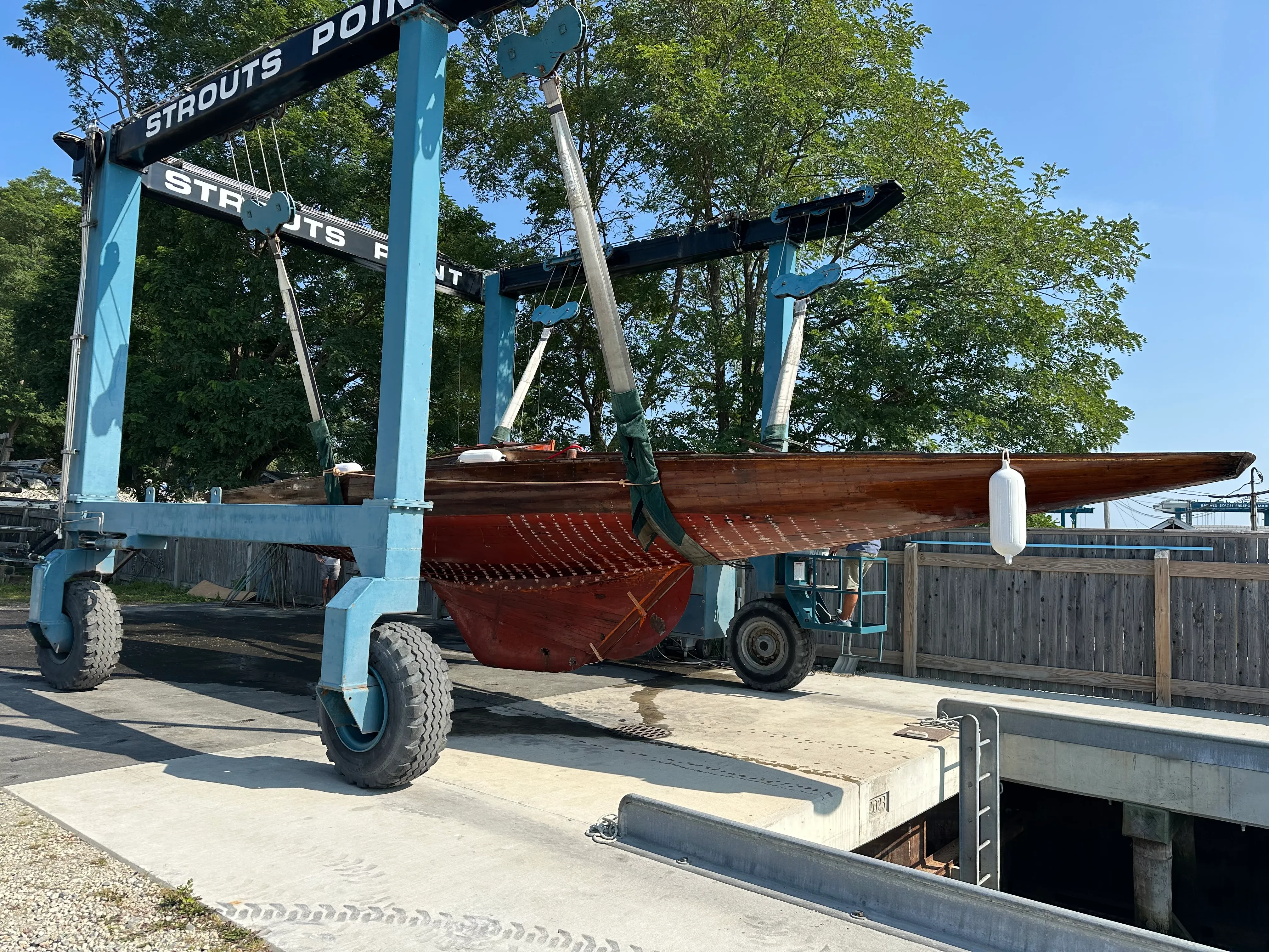 The sailboat suspended in the straps of a blue Travelift crane marked Strouts Point