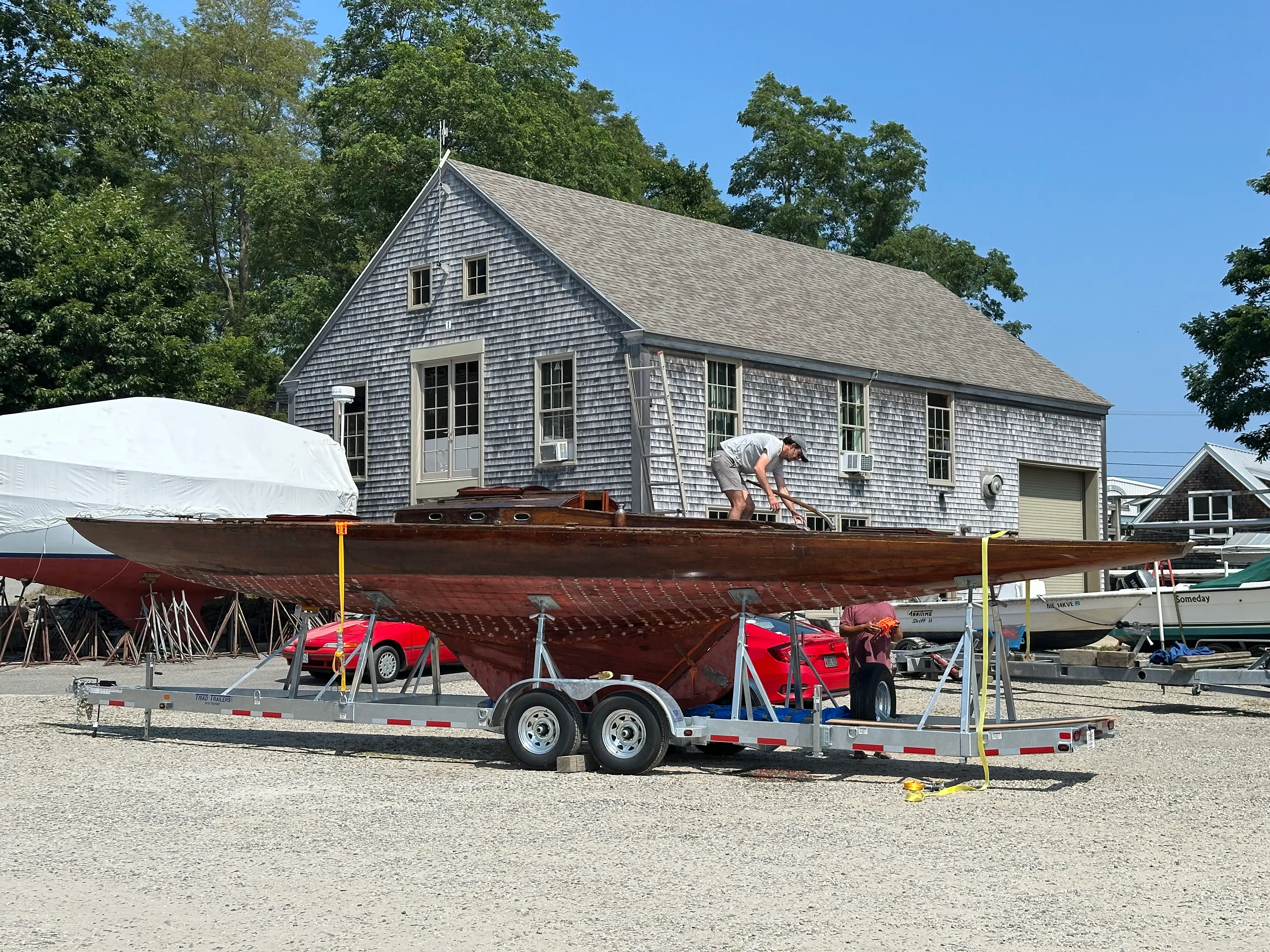 The sailboat on a flatbed trailer at the boatyard with a shingled boat shed behind