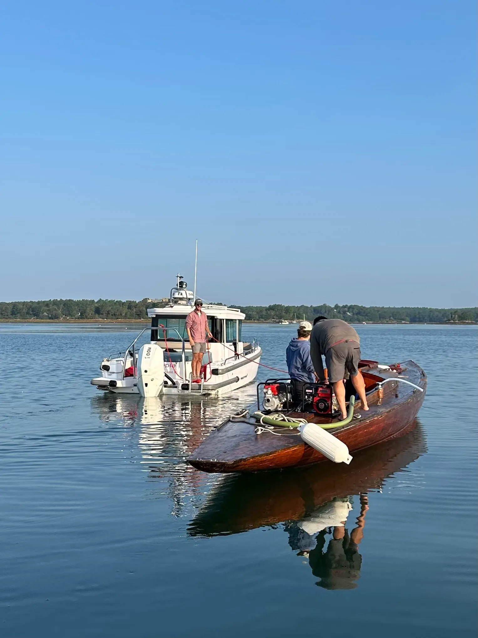 The wooden sailboat rafted alongside a white powerboat on open water, being towed